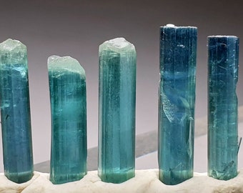 Puede incluir: Cinco cristales de aguamarina azul verdoso, cada uno con una forma y un tamaño diferentes, se muestran sobre una superficie blanca.