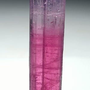 Puede incluir: Un cristal vertical y alargado con un degradado de tonos rosas, que va de un magenta intenso en la base a un rosa más claro y translúcido en la parte superior. La superficie del cristal presenta estrías visibles.