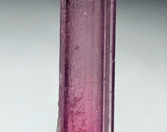 May include: A vertical, elongated pink tourmaline crystal. The crystal is a vibrant shade of pink, with a translucent quality. The surface has vertical striations and some minor imperfections. The crystal is mounted on a light-colored base.