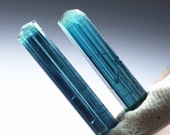 Puede incluir: Dos cristales largos, delgados, de color azul verdoso con estrías. Los cristales son translúcidos y tienen un brillo ligeramente iridiscente.