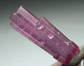 May include: A pink tourmaline crystal with a striated, faceted surface. The crystal is translucent and has a slight purple hue. The crystal is mounted on a gray base.