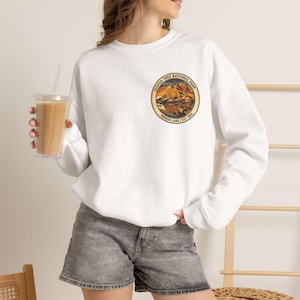 May include: A white long-sleeve sweatshirt featuring a circular graphic on the left chest. The graphic depicts a desert landscape with rock formations and water, labelled Joshua Tree National Park, Mirror Lake, and Est. 1994 in black text.