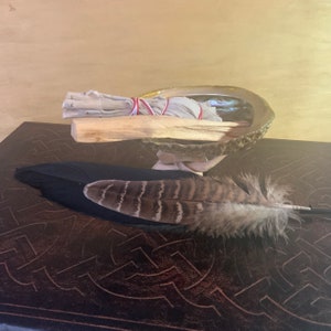 May include: A white sage smudge stick tied with red string, a wooden stick, and a brown and black turkey feather are arranged on a brown abalone shell. The shell is sitting on a dark brown surface with a Celtic knot design.