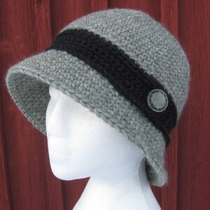 May include: A gray crocheted hat with a black band and a silver button. The hat is on a white mannequin head.