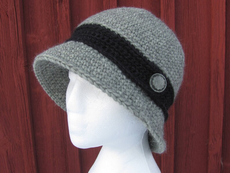 May include: A gray crocheted hat with a black band and a silver button. The hat is on a white mannequin head.