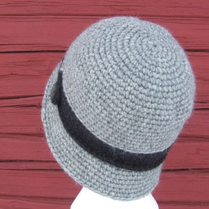 May include: A gray crocheted hat with a black band around the brim. The hat is sitting on a red wooden surface.