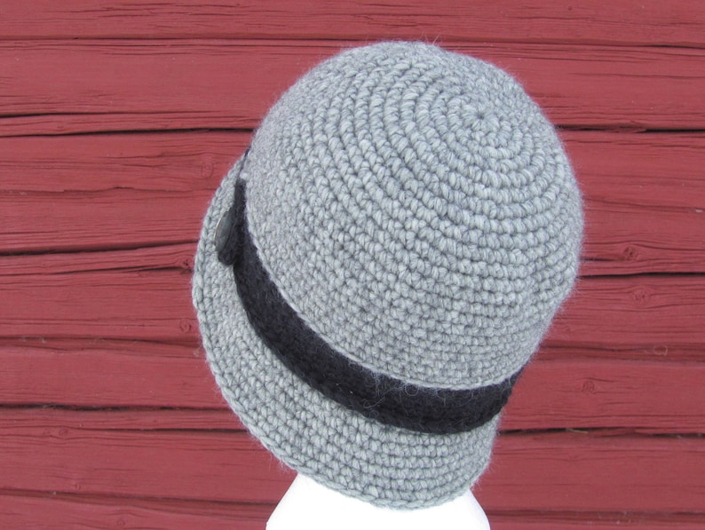 May include: A gray crocheted hat with a black band around the brim. The hat is sitting on a red wooden surface.