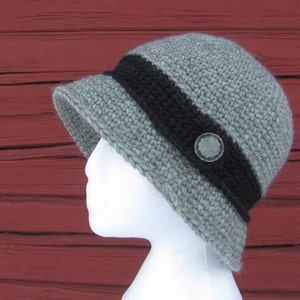 May include: A gray crocheted hat with a black band and a button. The hat is on a mannequin head against a red wooden background.