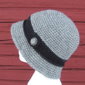 May include: A gray crocheted hat with a black band and a button. The hat is on a red wooden background.