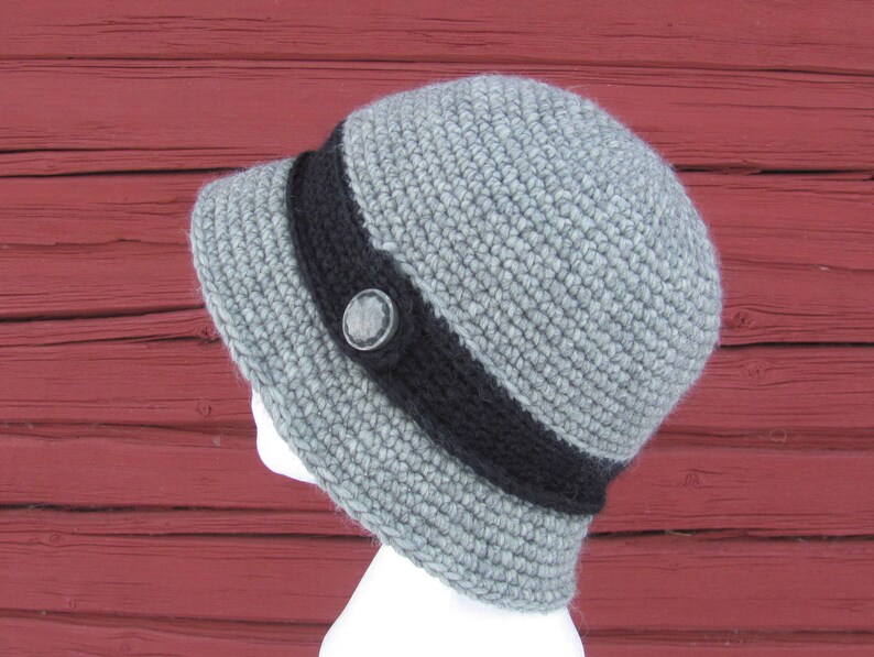 May include: A gray crocheted hat with a black band and a button. The hat is on a red wooden background.
