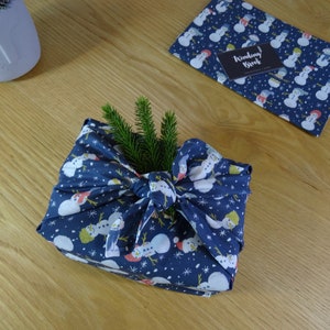 May include: A blue fabric gift wrap with a snowman pattern is tied with a bow and decorated with a sprig of green pine. The fabric has a white background with snowflakes and the text "Winding Brook" is visible on the fabric.