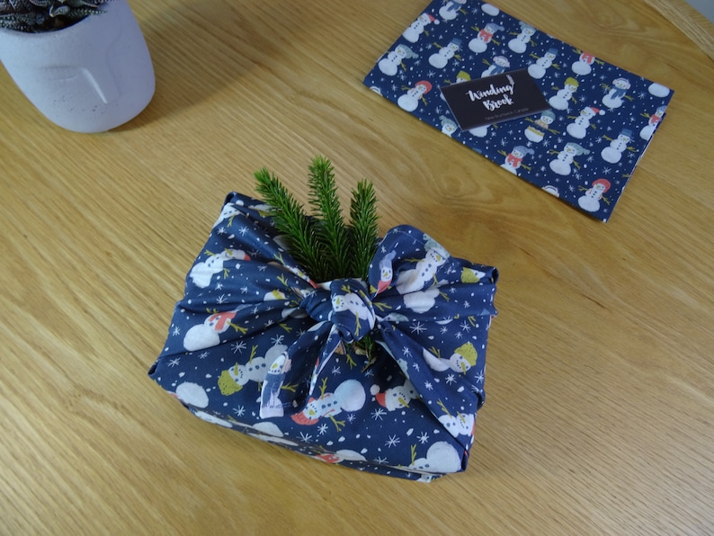 May include: A blue fabric gift wrap with a snowman pattern is tied with a bow and decorated with a sprig of green pine. The fabric has a white background with snowflakes and the text "Winding Brook" is visible on the fabric.