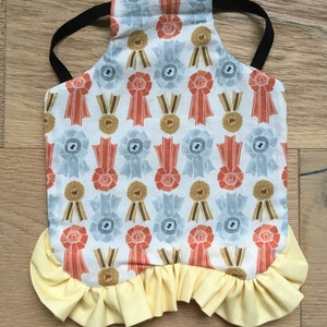 May include: A pet apron with a white background, decorated with repeating orange, blue, and gold ribbon designs. The apron has a yellow ruffled hem and a black elastic strap for securing. This pet accessory is designed for style and functionality.