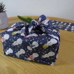 May include: A blue fabric gift wrap with a snowman pattern. The fabric is tied with a knot and a sprig of greenery is tucked into the knot.