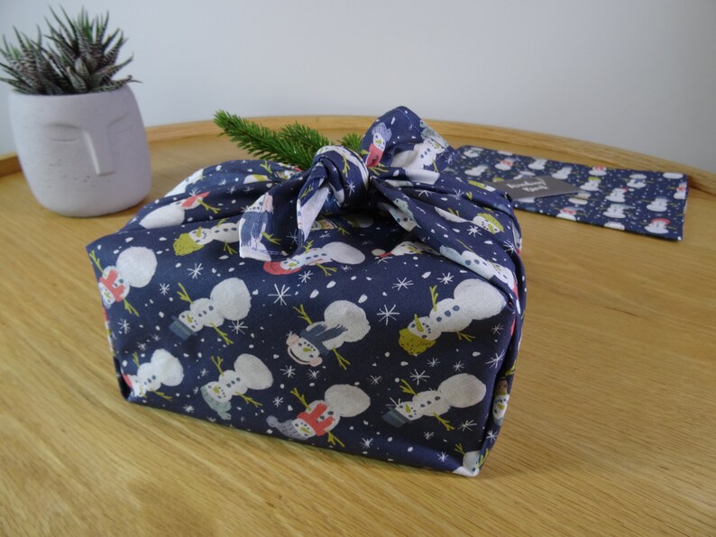May include: A blue fabric gift wrap with a snowman pattern. The fabric is tied with a knot and a sprig of greenery is tucked into the knot.