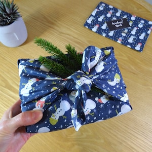 May include: A blue fabric gift wrap with a snowman pattern and white snowflakes. The fabric is tied with a bow and a sprig of greenery is placed on top.