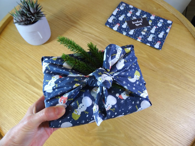 May include: A blue fabric gift wrap with a snowman pattern and white snowflakes. The fabric is tied with a bow and a sprig of greenery is placed on top.
