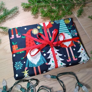 May include: A wrapped gift box with a black background and a festive pattern of gnomes, Christmas trees, and snowflakes. The box is tied with a red ribbon.