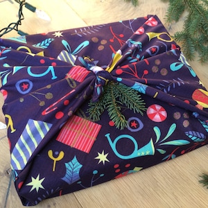 May include: A Christmas-themed fabric gift wrap with a colorful pattern of stars, holly, presents, and candy canes. The fabric is tied with a knot and decorated with a sprig of evergreen.