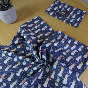 May include: A blue fabric with a white snowman pattern. The fabric is folded and shows the pattern on both sides. The fabric is on a wooden surface.