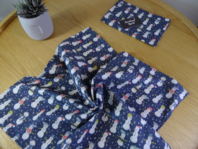 May include: A blue fabric with a white snowman pattern. The fabric is folded and shows the pattern on both sides. The fabric is on a wooden surface.