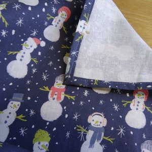 May include: A navy blue fabric with a white background featuring a repeating pattern of snowmen with different hats and scarves. The fabric has a white border around the edges.