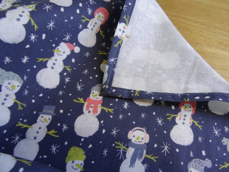 May include: A navy blue fabric with a white background featuring a repeating pattern of snowmen with different hats and scarves. The fabric has a white border around the edges.