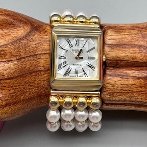 May include: A gold quartz watch with a rectangular face and a pearl bracelet. The watch face is white with black Roman numerals and hands. The bracelet is made of multiple strands of white pearls.