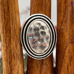 May include: Silver-toned ring with an oval, hammered-metal center. The ring has a double black band around the hammered center. The ring is displayed on a wooden ring holder.