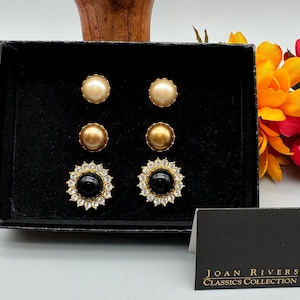 May include: A set of three pairs of earrings in a black box. The earrings are gold-toned and feature a pearl, a gold ball, and a black stone surrounded by clear rhinestones. The box has a black label that reads "JOAN RIVERS CLASSICS COLLECTION".