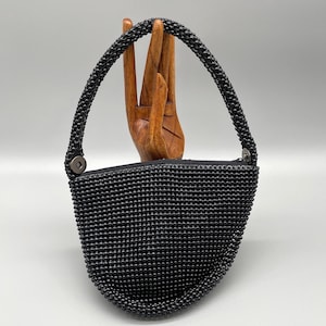 May include: A small, black beaded handbag with a rounded bottom and a zipper closure. The handle is also beaded and attached to the bag with metal hardware. The bag is displayed against a neutral background.