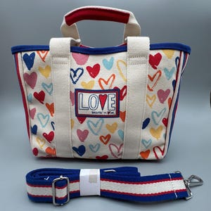Puede incluir: Un bolso tote de lona con un estampado de corazones coloridos y la palabra "LOVE" en la parte delantera. El bolso tiene un ribete rojo y azul, asas blancas y una correa de hombro a rayas azul y blanca desmontable.