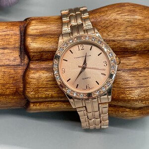 May include: A rose gold wristwatch with a rose gold band and a white face. The face has a rose gold trim and is adorned with small clear crystals. The watch face has black hands and black numbers. The watch face also has the words 'Charter Club' and 'Quartz' printed on it. The watch face also has the words 'China Mov't' printed on it.