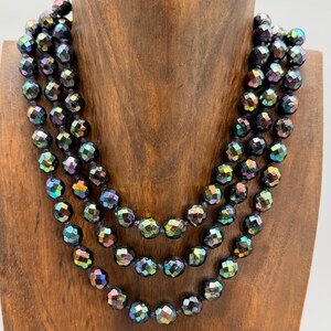 Puede incluir: Un collar de tres hilos con cuentas facetadas e iridiscentes. Las cuentas son principalmente negras con toques de verde, azul y morado. El collar se exhibe en un busto de madera, mostrando su diseño en capas y variaciones de color.