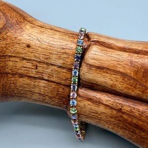 Puede incluir: Una pulsera dorada con una fila de gemas multicolores. Las gemas son de color rosa, azul, verde y morado. La pulsera está sobre un trozo de madera con un fondo azul claro.