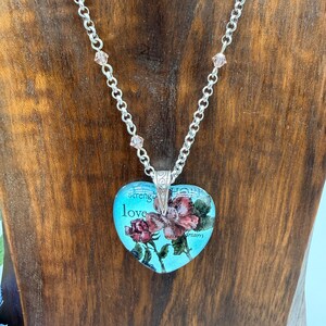May include: A heart-shaped pendant necklace with a silver chain and pink crystal accents. The pendant features a floral design with the words "strength", "love", and "dream" on a turquoise background.