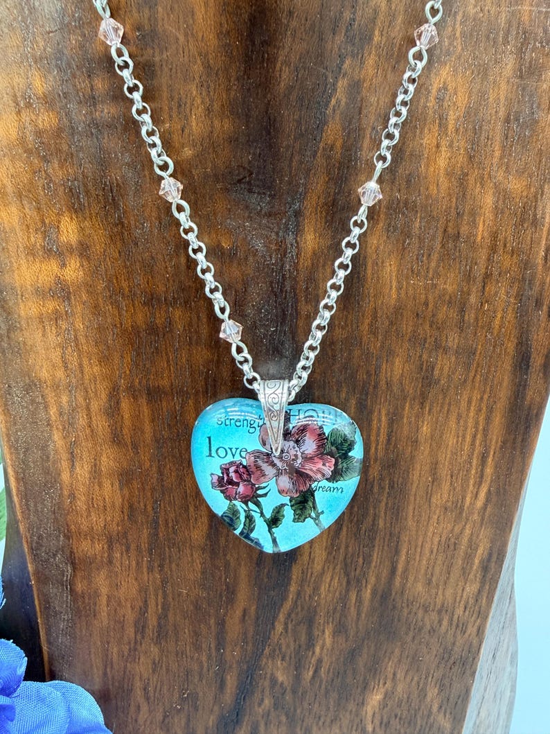 May include: A heart-shaped pendant necklace with a silver chain and pink crystal accents. The pendant features a floral design with the words "strength", "love", and "dream" on a turquoise background.
