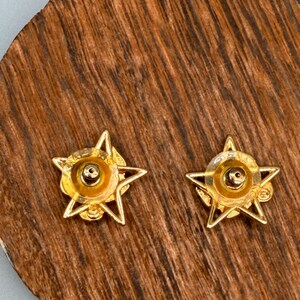 May include: A pair of gold-colored star-shaped stud earrings. Each earring features a five-pointed star design with a central circular element. The earrings are displayed on a wooden surface with a natural grain pattern.