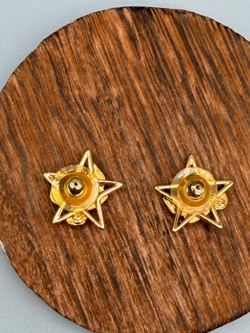 May include: A pair of gold-colored star-shaped stud earrings. Each earring features a five-pointed star design with a central circular element. The earrings are displayed on a wooden surface with a natural grain pattern.