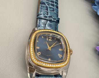 May include: A wristwatch with a blue crocodile-textured leather strap, a clear plastic case, and a gold-tone bezel encrusted with small clear stones. The dark blue dial features gold-tone hands, markers, and the text Joan Rivers Quartz.