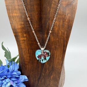 May include: A silver-toned necklace with a heart-shaped pendant. The pendant features a floral design with the words "strength" and "love" on a light blue background. The necklace is displayed on a wooden stand with a blue flower in the foreground.