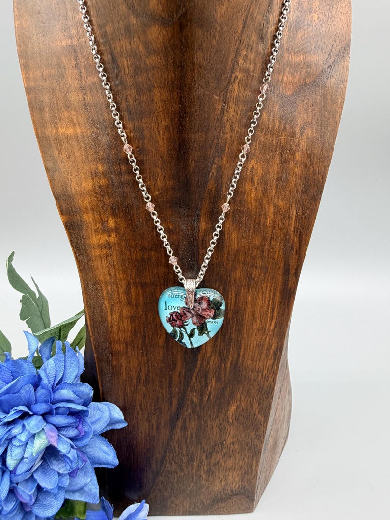 May include: A silver-toned necklace with a heart-shaped pendant. The pendant features a floral design with the words "strength" and "love" on a light blue background. The necklace is displayed on a wooden stand with a blue flower in the foreground.