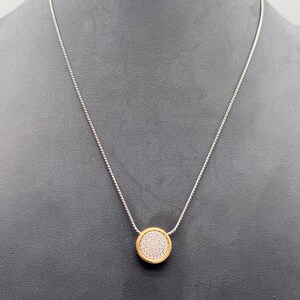 Puede incluir: Collar de cadena plateada con un colgante circular dorado. El colgante tiene un centro texturizado y de color claro. El collar se muestra sobre un fondo oscuro.