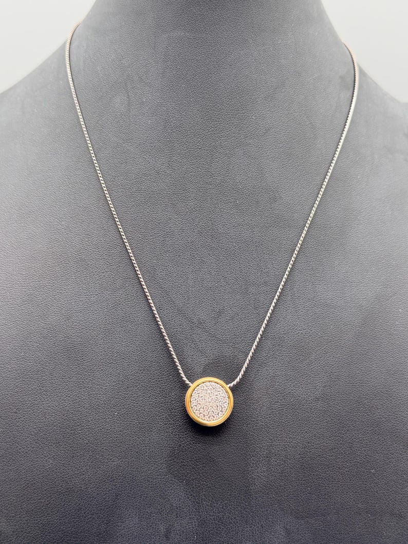 Puede incluir: Collar de cadena plateada con un colgante circular dorado. El colgante tiene un centro texturizado y de color claro. El collar se muestra sobre un fondo oscuro.