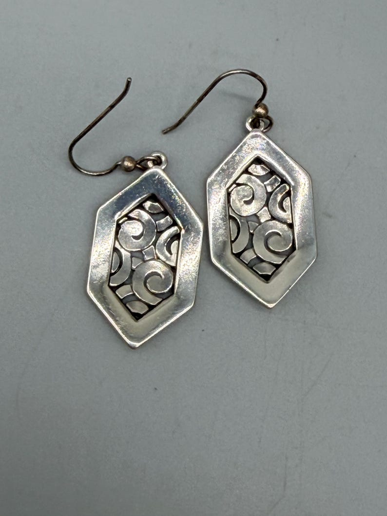 Puede incluir: Un par de pendientes plateados. Cada pendiente presenta un marco geom&eacute;trico hexagonal con un dise&ntilde;o calado de espirales. Los pendientes tienen un cierre de gancho y una peque&ntilde;a cuenta dorada cerca de la parte superior.