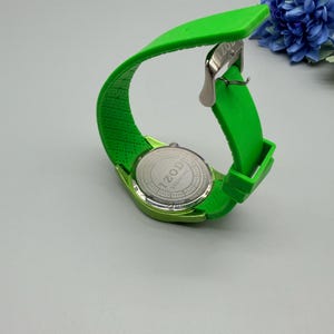 May include: A bright lime green wristwatch with a flexible silicone strap and a silver-tone metal buckle. The back of the watch case is stainless steel and features the engraved text IZOD ESSENTIAL and JAPAN MOVEMENT.