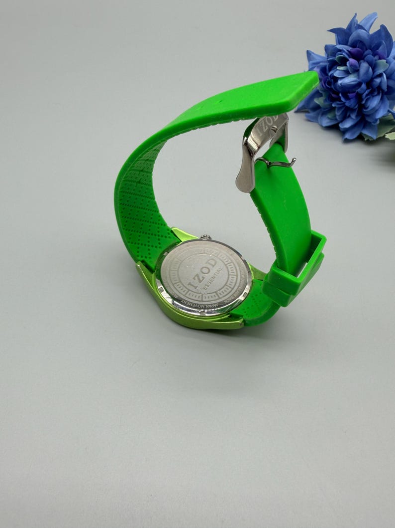 May include: A bright lime green wristwatch with a flexible silicone strap and a silver-tone metal buckle. The back of the watch case is stainless steel and features the engraved text IZOD ESSENTIAL and JAPAN MOVEMENT.