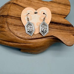 Puede incluir: Un par de pendientes geom&eacute;tricos plateados con un dise&ntilde;o de filigrana y un borde de peque&ntilde;as piedras transparentes. Los pendientes se exhiben en una etiqueta en forma de coraz&oacute;n sobre un fondo de madera.