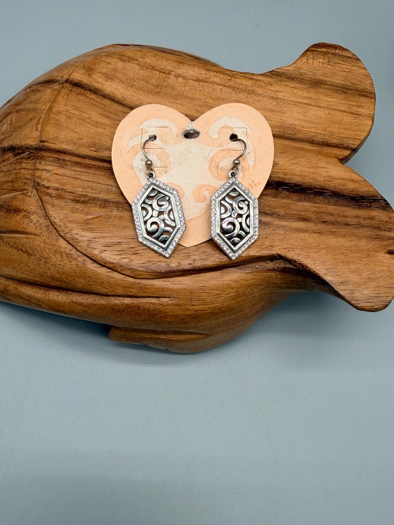 Puede incluir: Un par de pendientes geom&eacute;tricos plateados con un dise&ntilde;o de filigrana y un borde de peque&ntilde;as piedras transparentes. Los pendientes se exhiben en una etiqueta en forma de coraz&oacute;n sobre un fondo de madera.