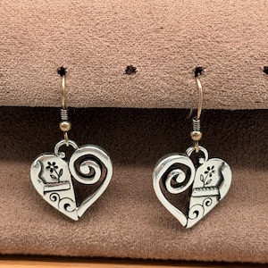 May include: A pair of silver-coloured heart-shaped earrings. Each earring has a floral design on one side and a swirling design on the other. The earrings hang from a gold-coloured hook.